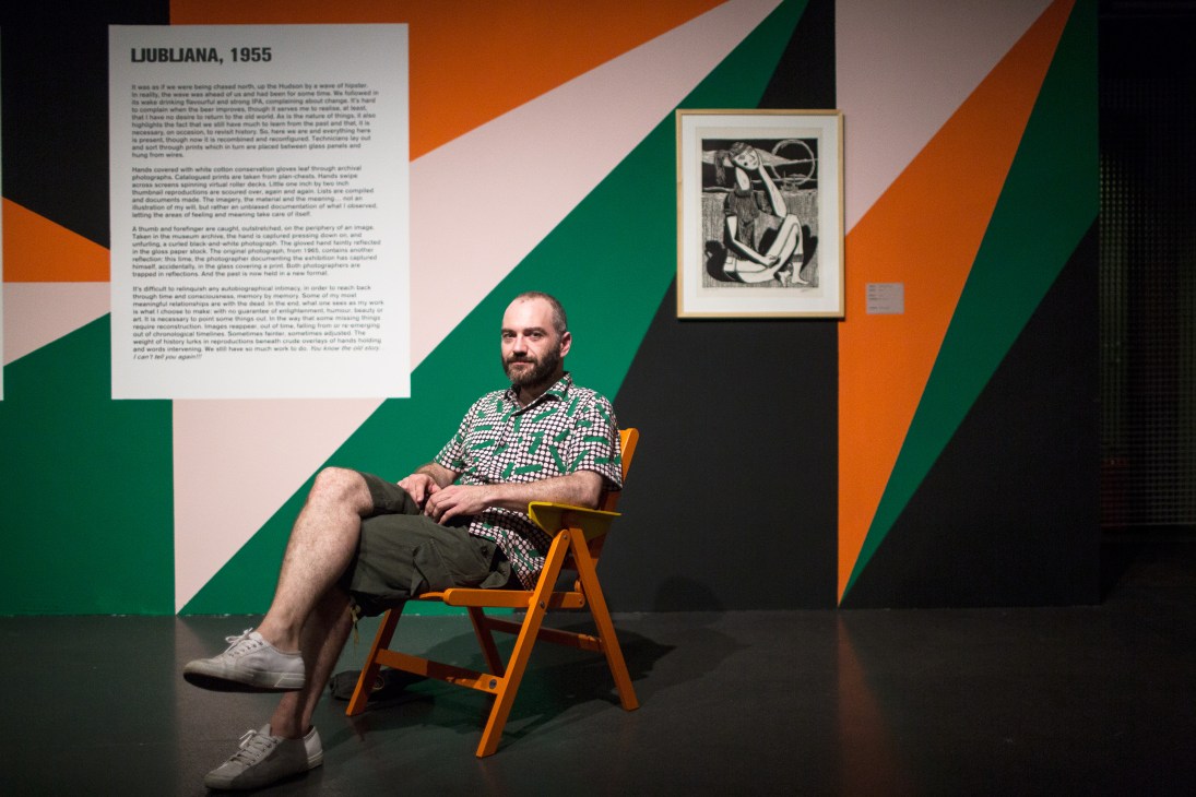 Artist Giles Round seated on an orange chair in front of colorful exhibition wall with graphic artwork displayed.
