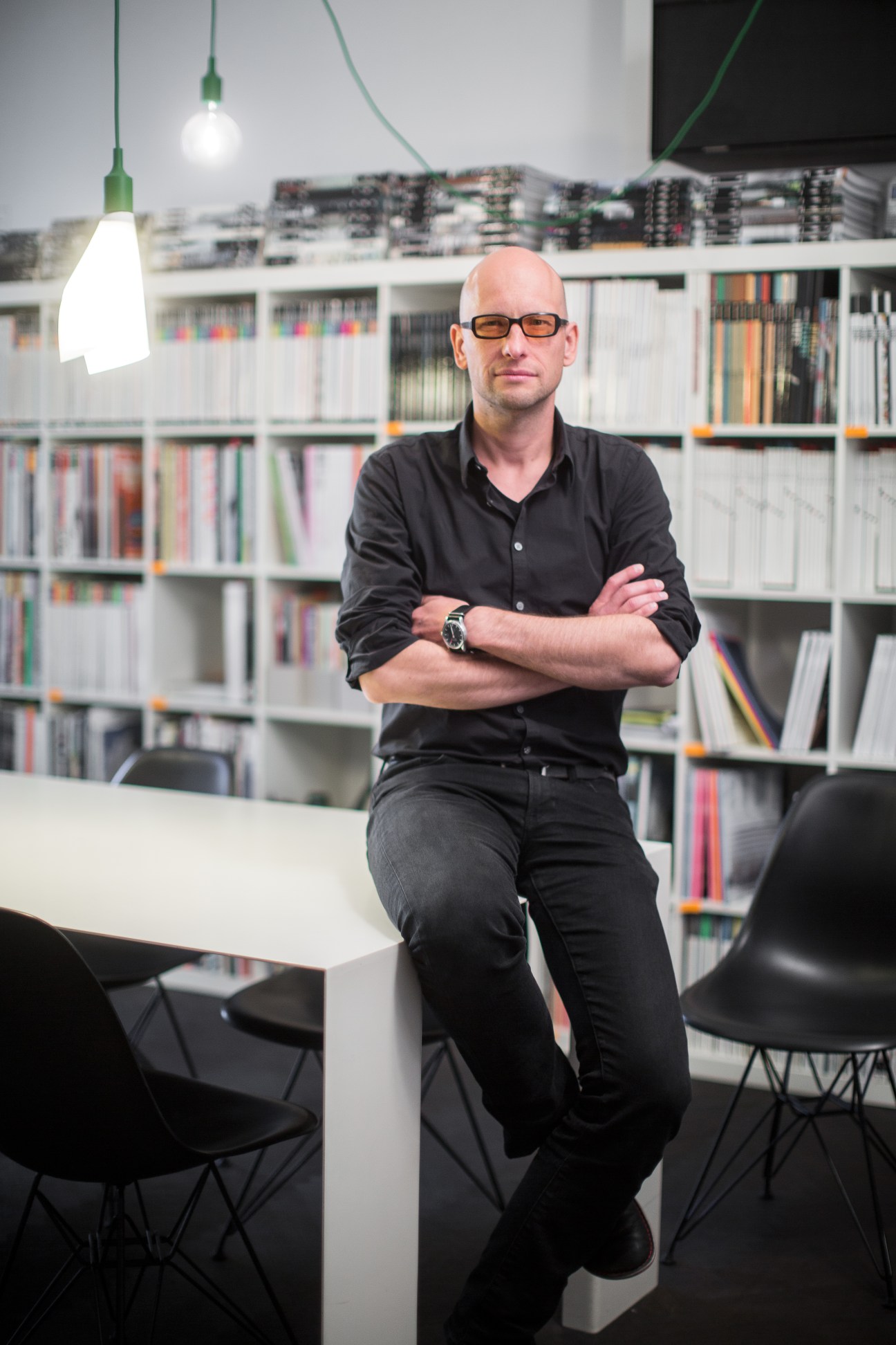 Architect Aljosa Dekleva sitting on a white table with arms crossed in an office with bookshelves and hanging pendant lights.