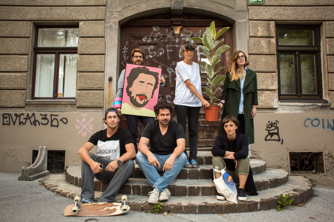 Six young designers pose on steps outside a graffitied building entrance, one holding a colorful portrait artwork.