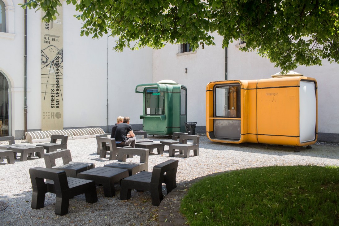 Green and yellow K67 kiosks in a Ljubljana courtyard with seating and signage.