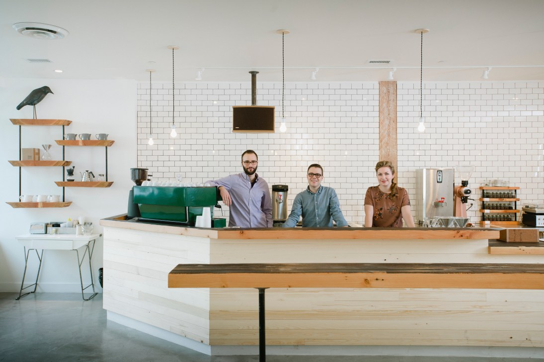 Three staff members stand behind the counter at Matchstick café in Vancouver, with white subway tiles and wooden shelving visible.