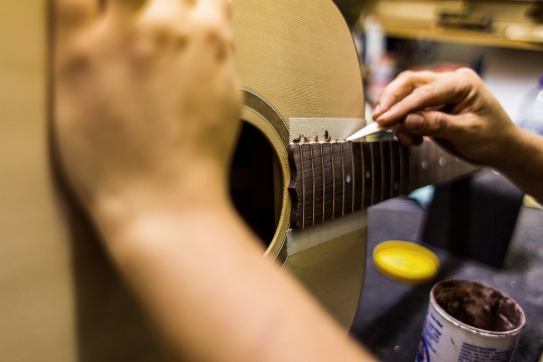 A craftsperson performs repair work on a Maton acoustic guitar, carefully working on the fretboard with a tool.