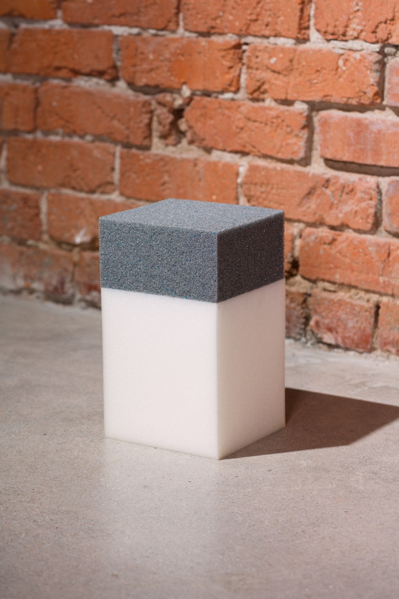 Cube-shaped foam sample with dark grey top half and white bottom half against a brick wall.