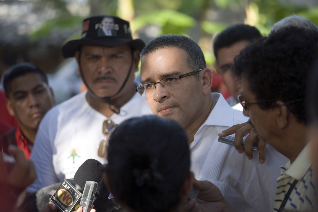 Mauricio Funes speaks to reporters at a press conference surrounded by microphones and supporters.