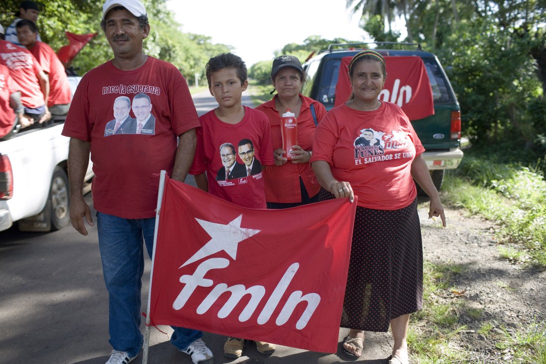 FMLN supporters wearing red campaign t-shirts hold a Funes campaign banner on a rural road.