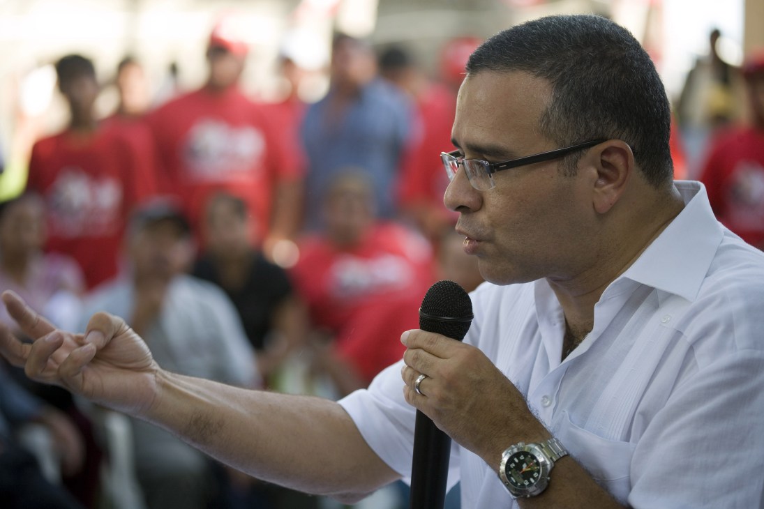 Mauricio Funes speaks to supporters holding a microphone, wearing glasses and a white shirt.