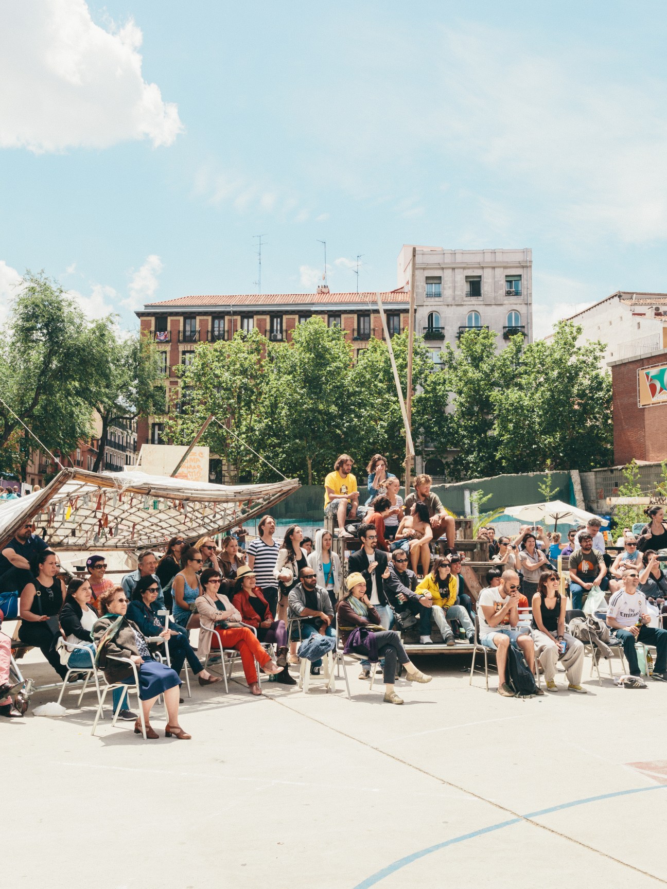 Community gathering at Campo de Cebada with people seated on chairs facing wooden structures and green trees in Madrid.