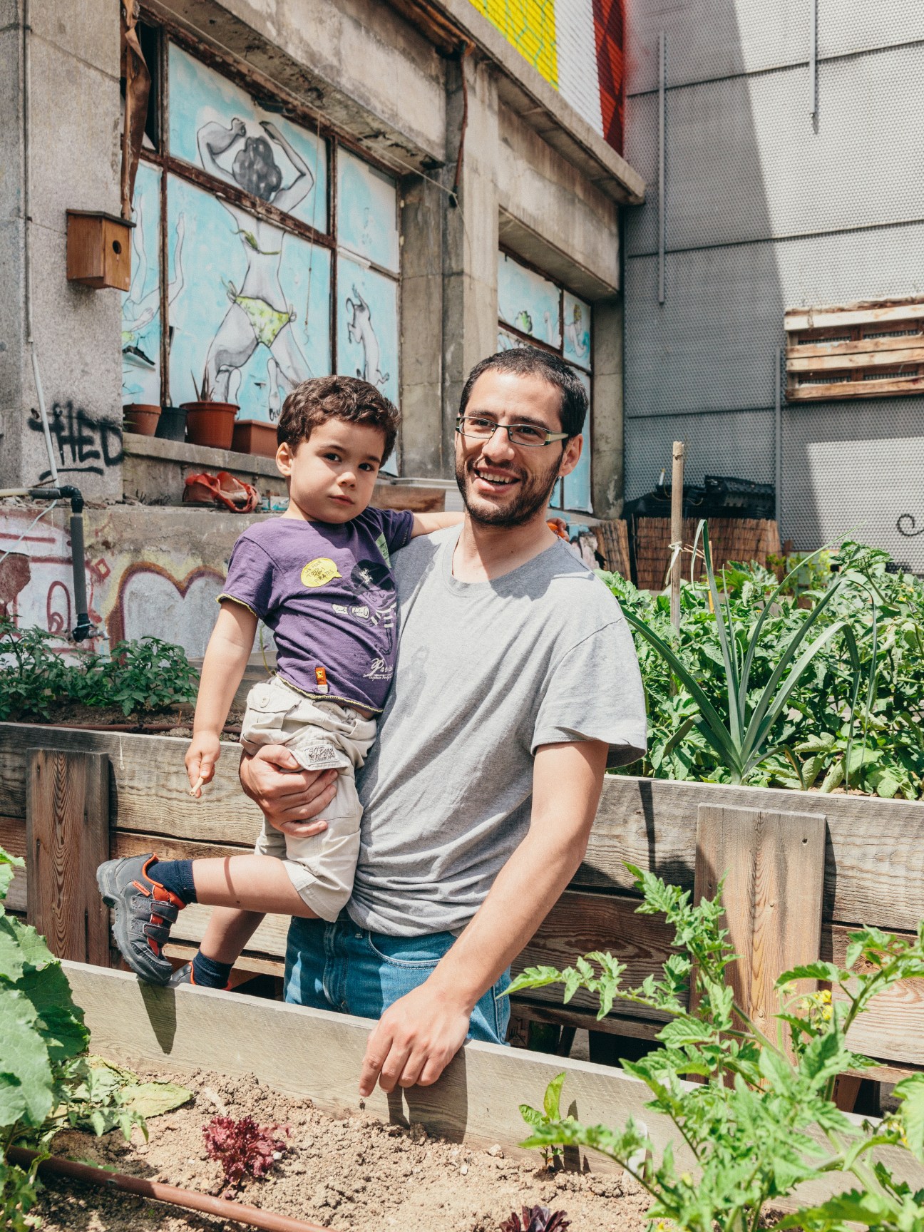 Man holding a young child in an urban garden with raised wooden planting beds and muraled building backdrop.