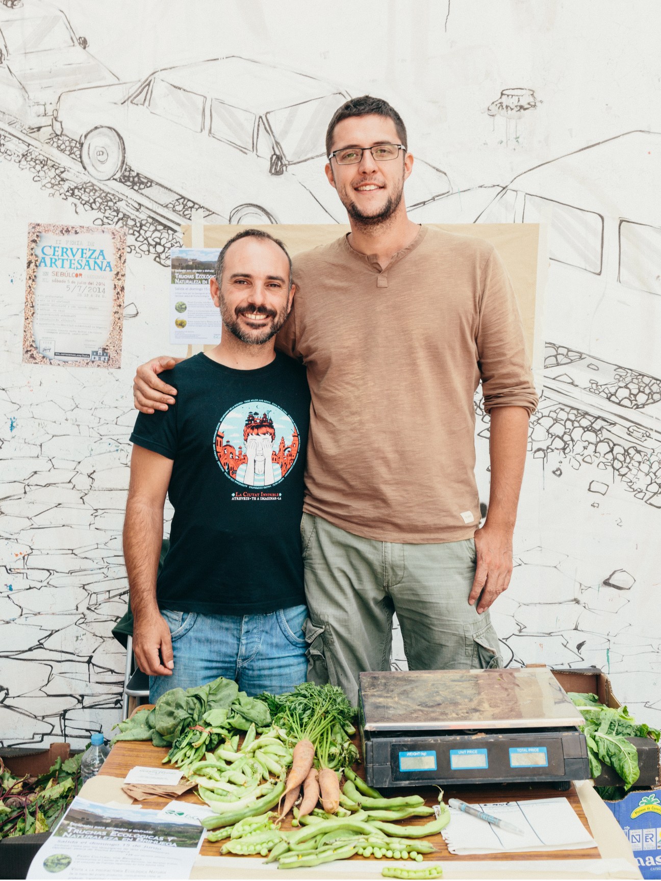 Two men stand together in front of a wall with architectural sketches, with fresh vegetables displayed on a table in front of them.