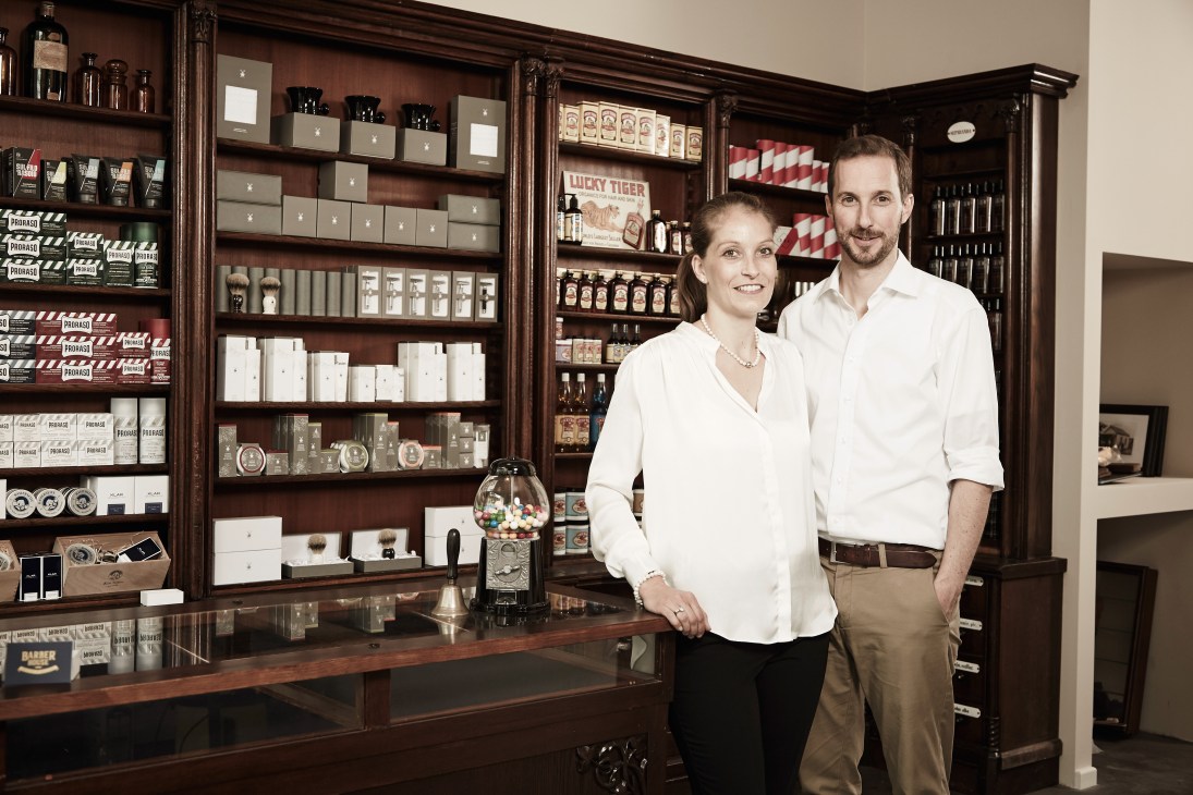 Two people in white shirts stand at a wooden counter inside Barber House barbershop in Munich, with shelves of grooming products displayed behind them.
