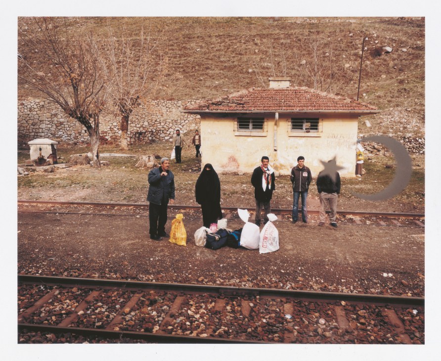 People standing by a small building next to railway tracks in a rural Turkish landscape.