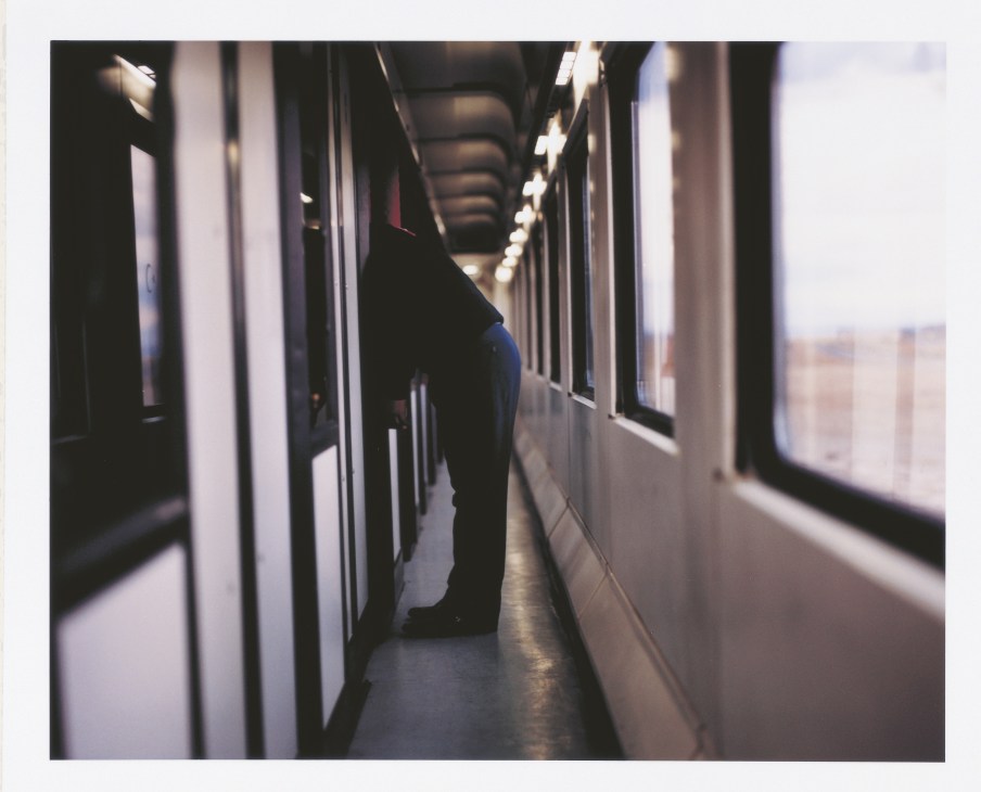 A passenger stands in a narrow, dimly lit corridor of the TransAsya Express train with windows lining one side.