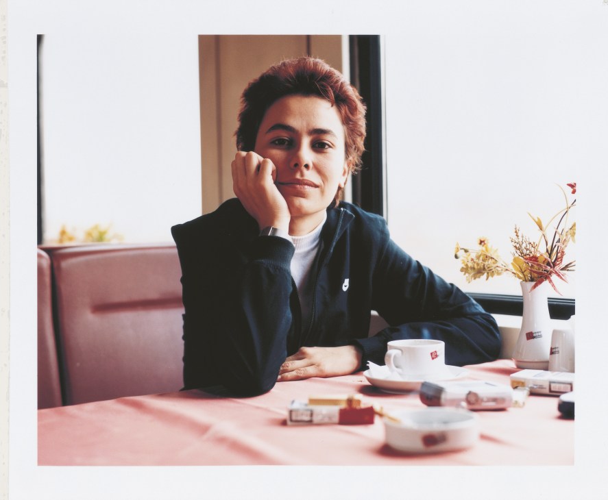 Woman with reddish hair in dark jacket sits at pink table with coffee cup, hand to face.