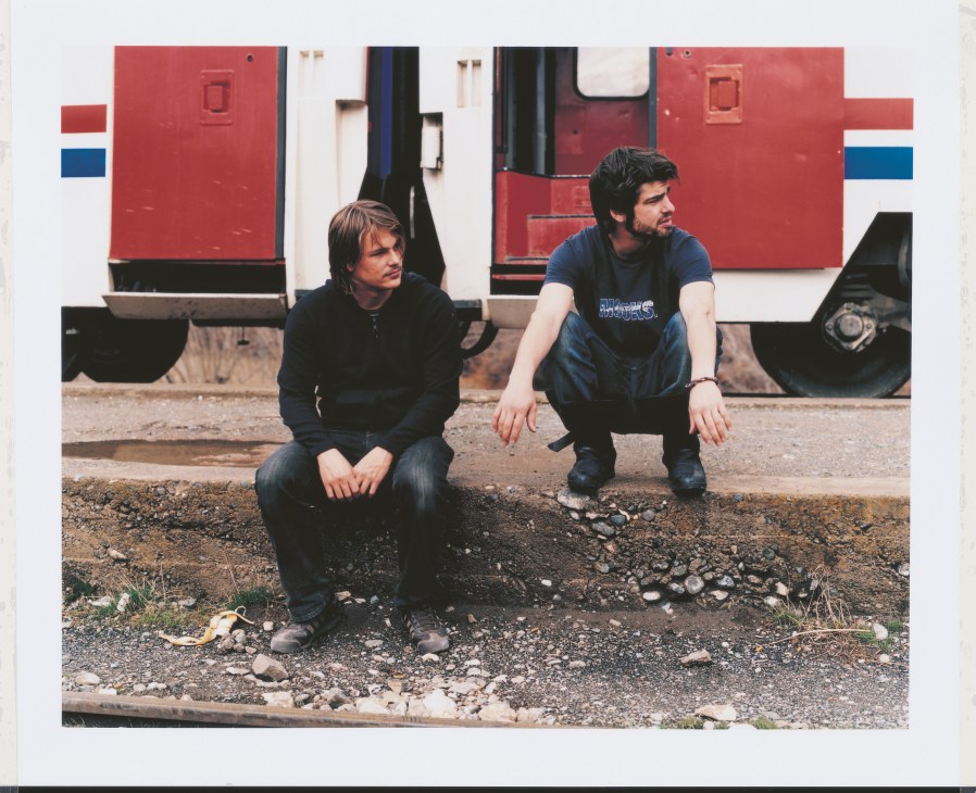 Two young men in casual clothing crouch in front of red and blue train cars.