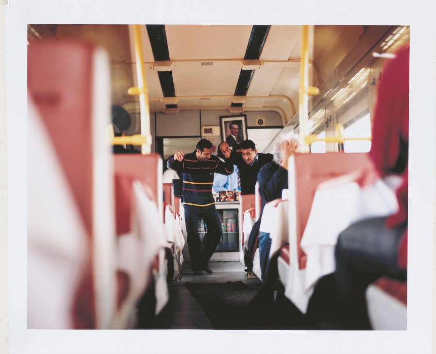 Two men dance in a train dining car with red seats and yellow handrails visible.