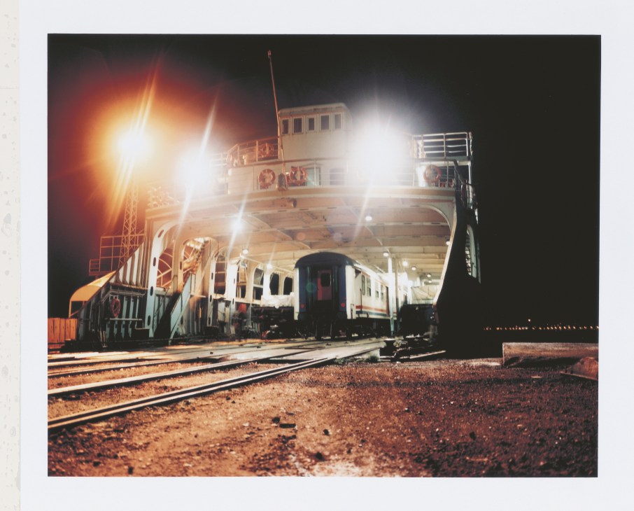Night scene of a ferry with train car visible, illuminated by bright lights at a dock.