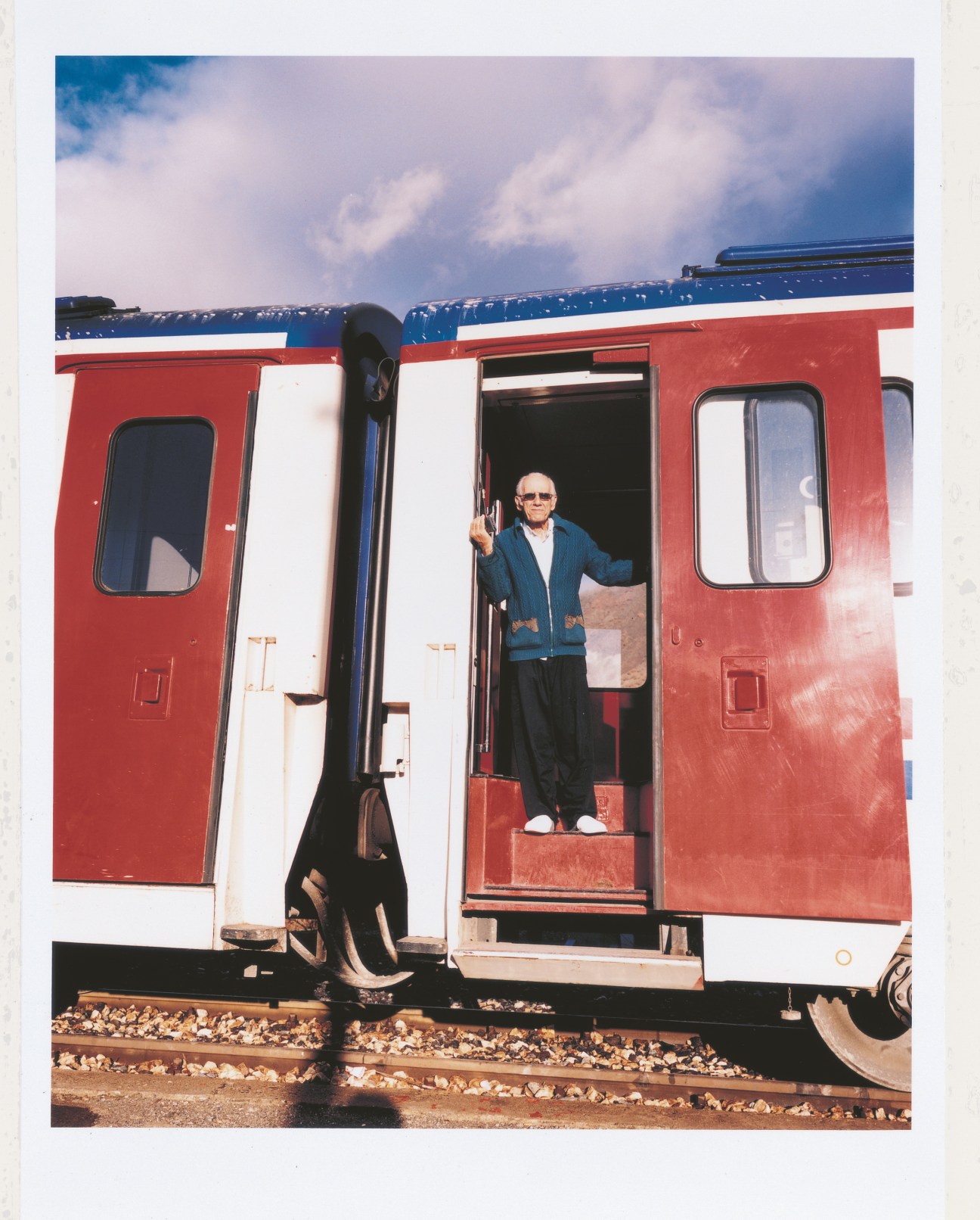 A man in sunglasses and blue jacket stands in the doorway of a red and white train carriage.