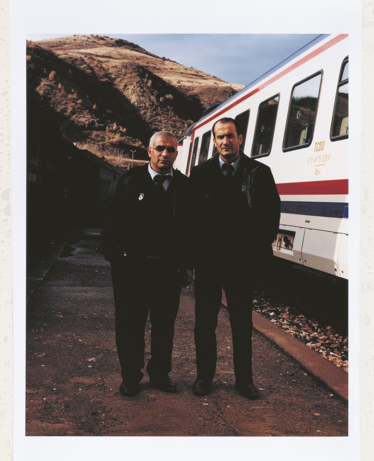 Two armed guards in dark suits stand outside a Turkish State Railways train with mountains behind them.