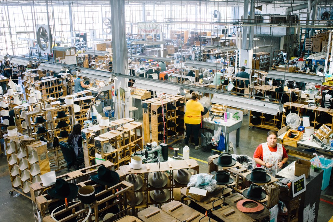 Workers at industrial stations assembling hats in a large, bright factory floor with wide windows and equipment.