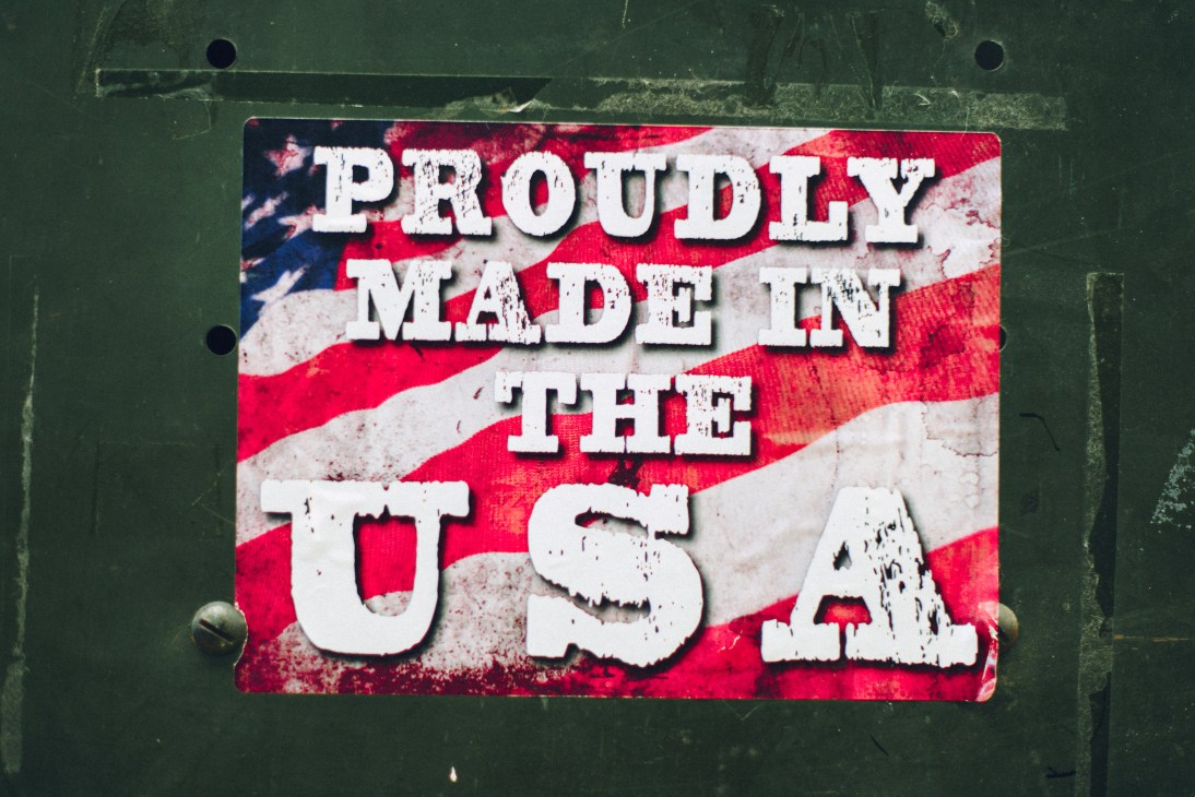 Weathered "Proudly Made in the USA" sign with American flag background mounted on green wall