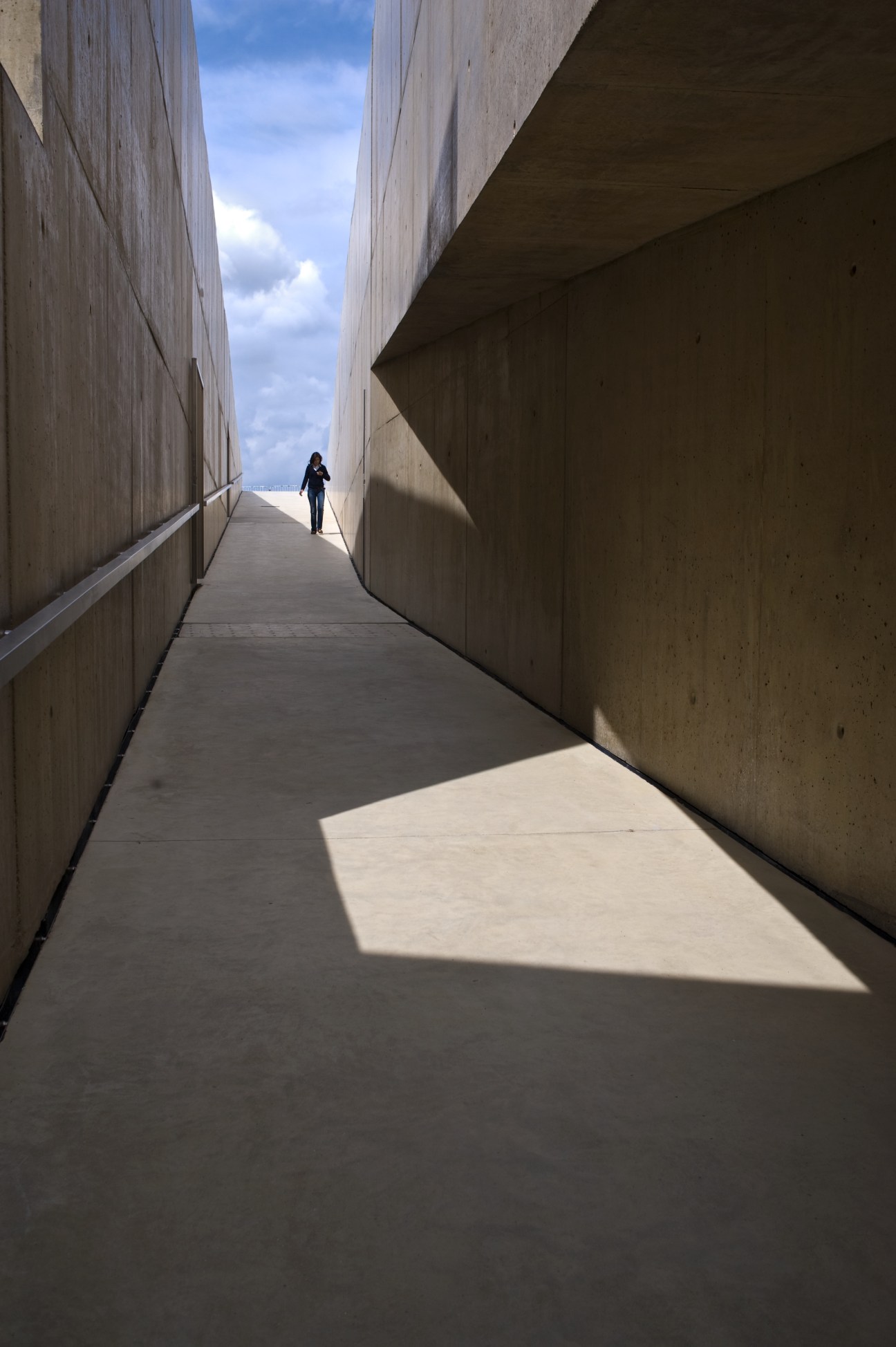 A person walks through a narrow concrete corridor with high walls and natural light streaming in from above.