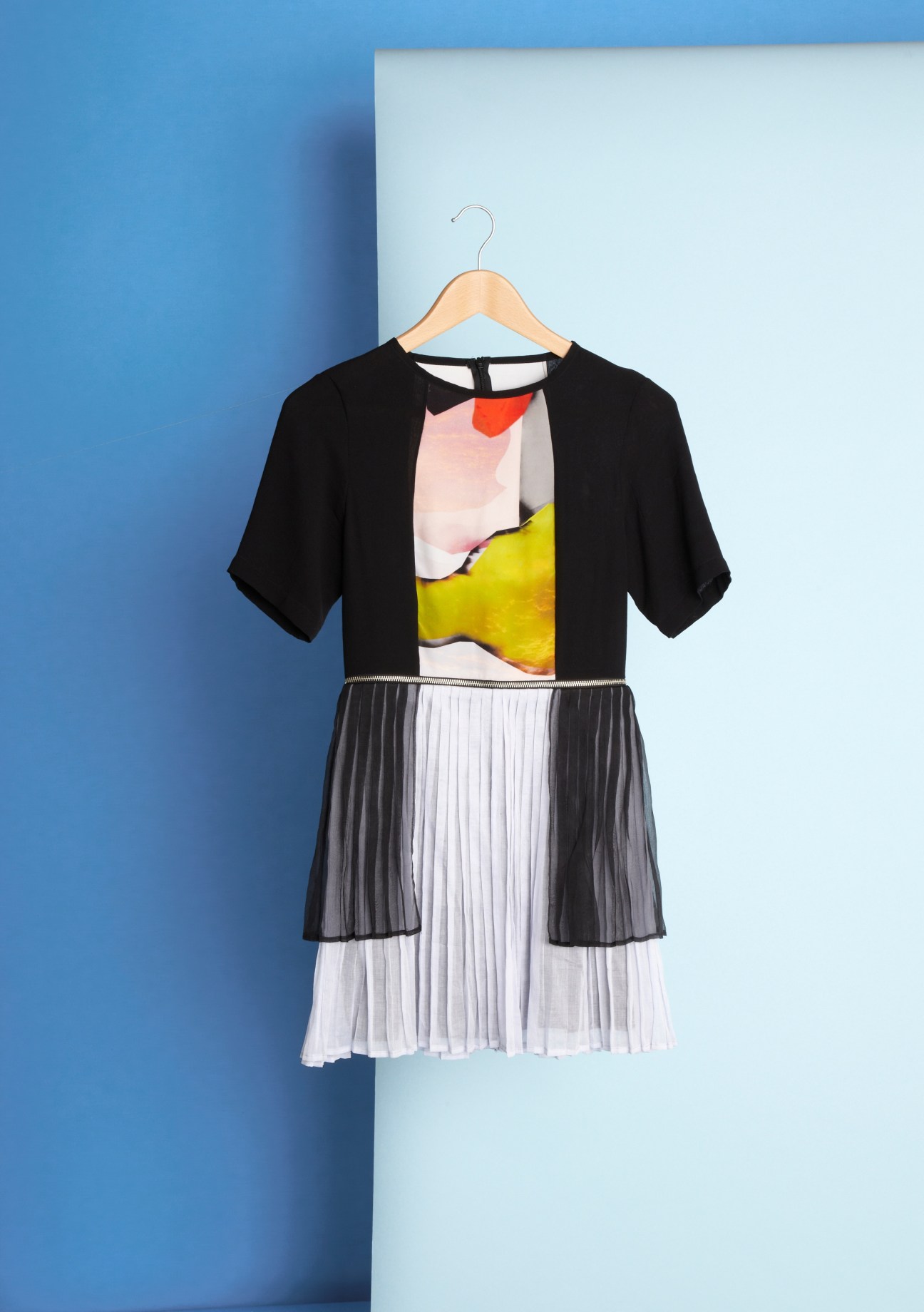 Black short-sleeve dress with colorful abstract print and pleated white skirt on wooden hanger against blue backdrop.