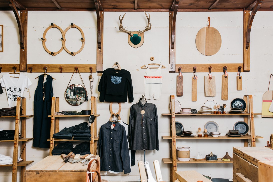 M Crow shop interior displaying clothing and homewares on wooden shelves against white shiplap walls.