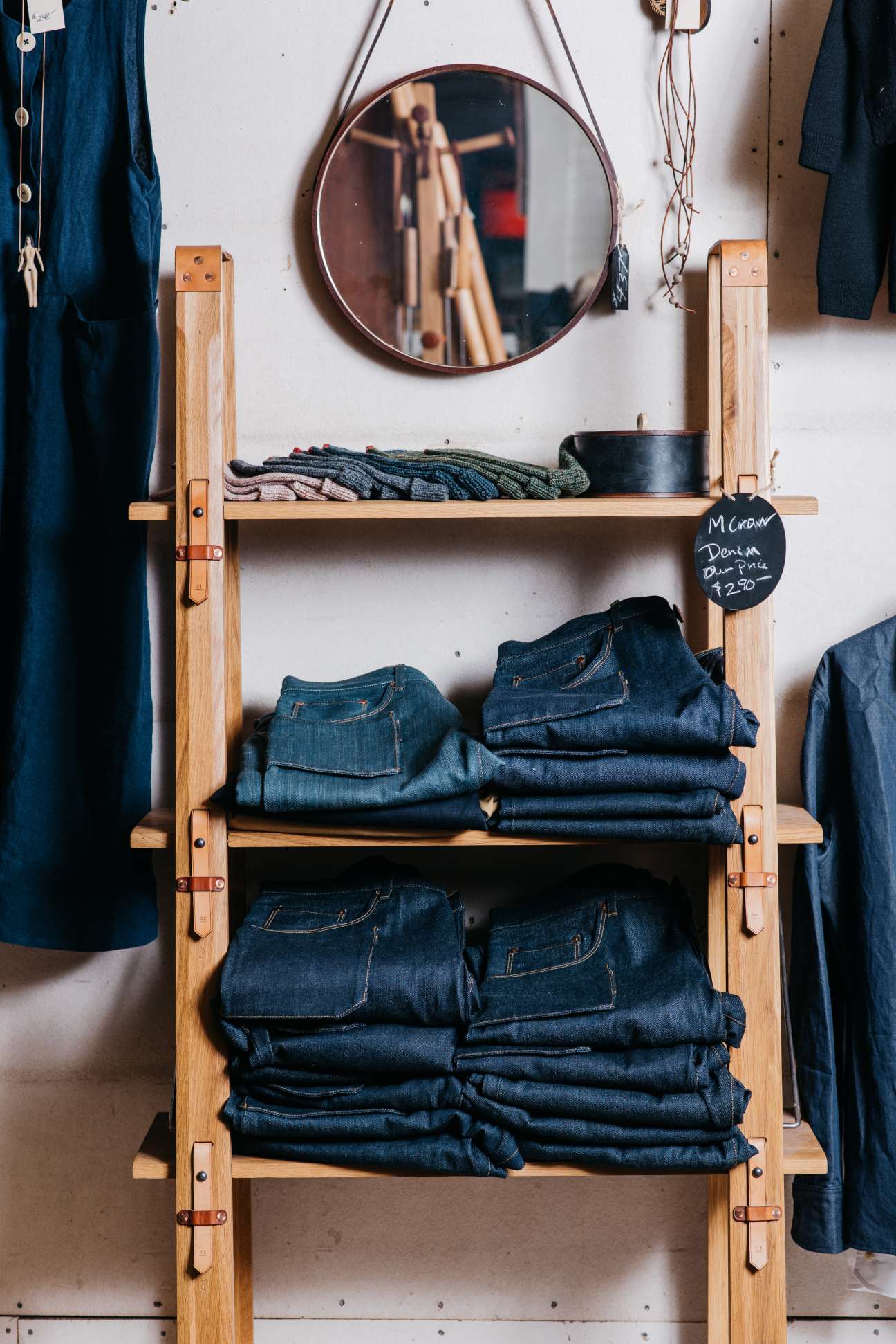 Wooden shelving unit displaying folded denim jeans and textiles with a round mirror and price tag for M Crow denim.