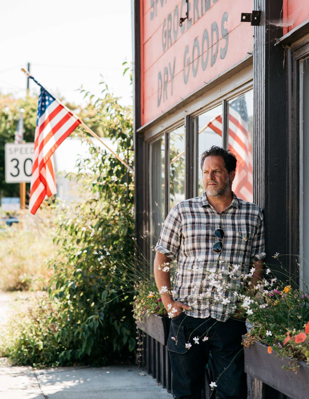 Tyler Hays stands outside M Crow general store wearing a plaid shirt with an American flag visible behind him.
