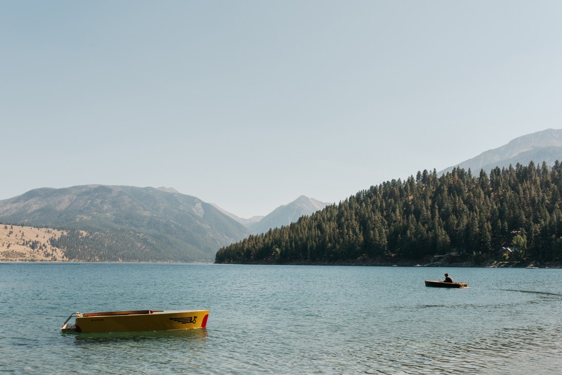 Boats float on turquoise water with forested mountains and clear sky in background.