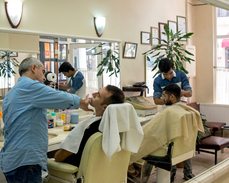 Huseyin Akbay and apprentice Aydin Simsek work on customers in a modern barbershop with mirrors and plants.