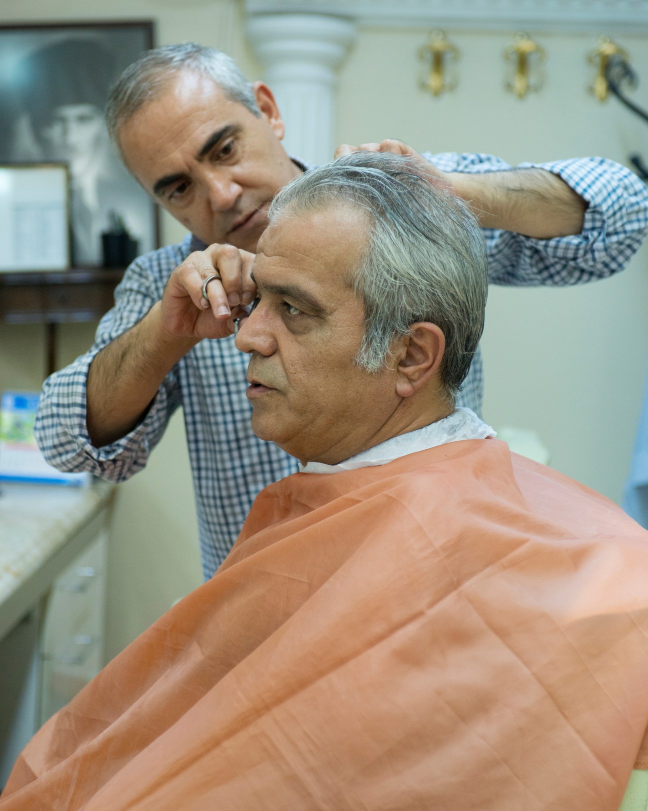 A barber in a blue checkered shirt cuts an older man's hair in a traditional Istanbul barbershop.