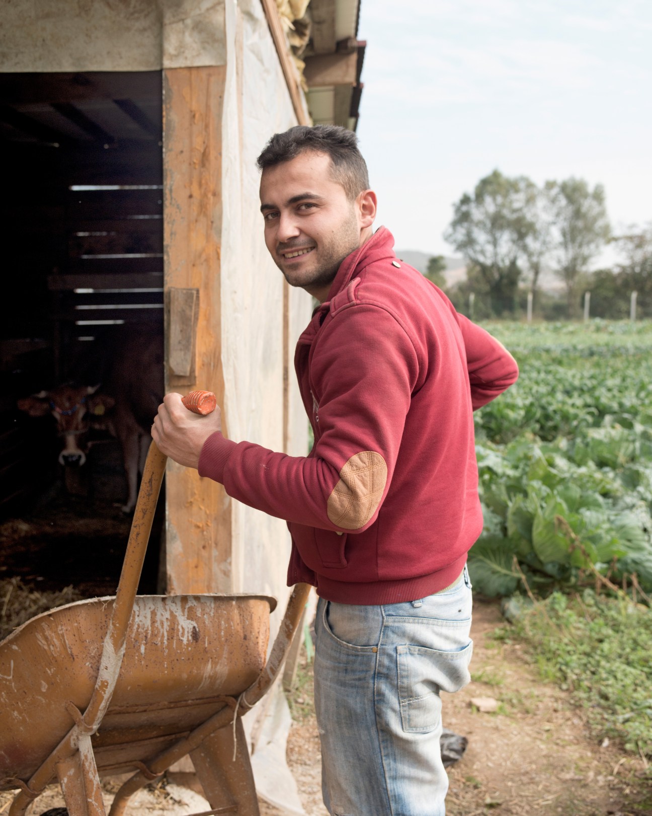 A man in a red hoodie holding a wooden pole outside a farm building with vegetable fields visible in the background.