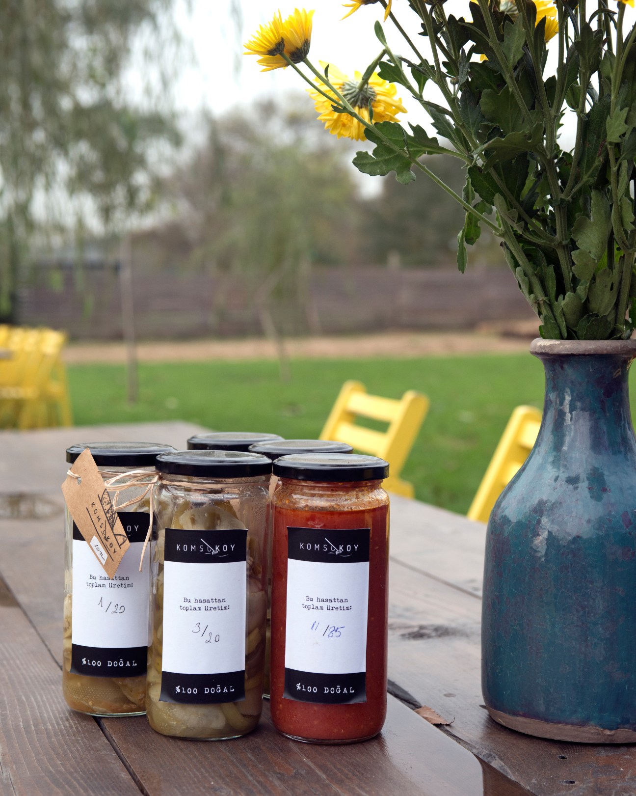 Jars of Komsukoy farm products with handwritten labels on outdoor table next to blue vase with yellow flowers.