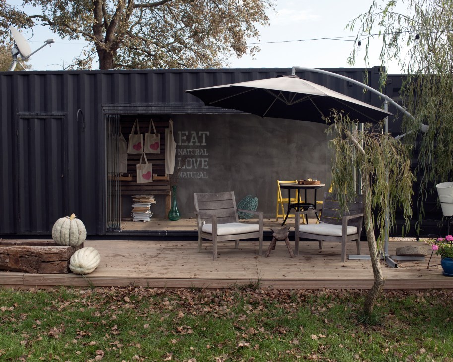Modern black shipping container cafe with outdoor seating, umbrella, and signage reading 'Eat Natural Love Natural'