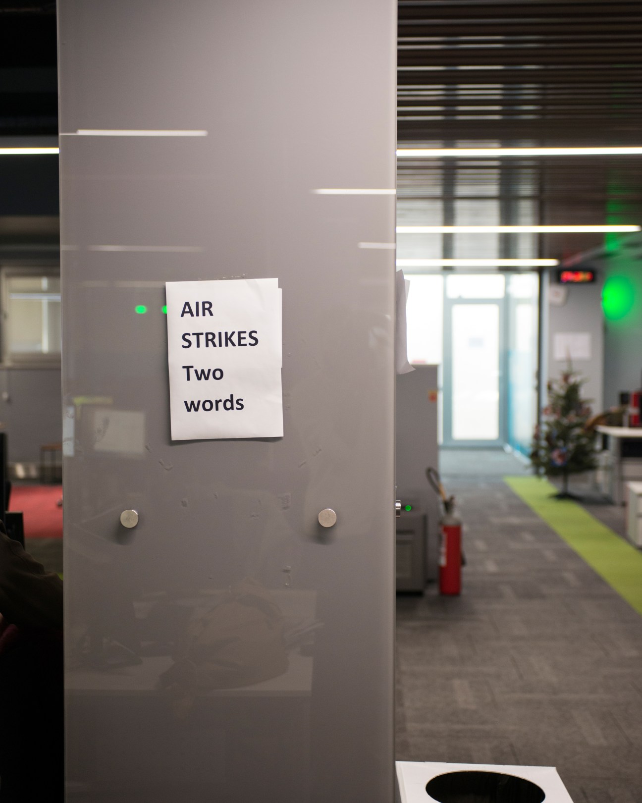 A white paper sign reading 'AIR STRIKES Two words' taped to a glass partition in a modern newsroom office.