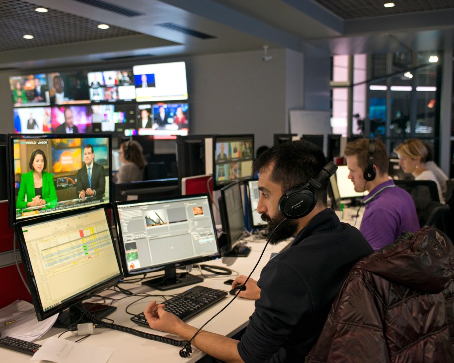 Newsroom staff working at computer stations with multiple monitors displaying broadcast feeds and editing software.
