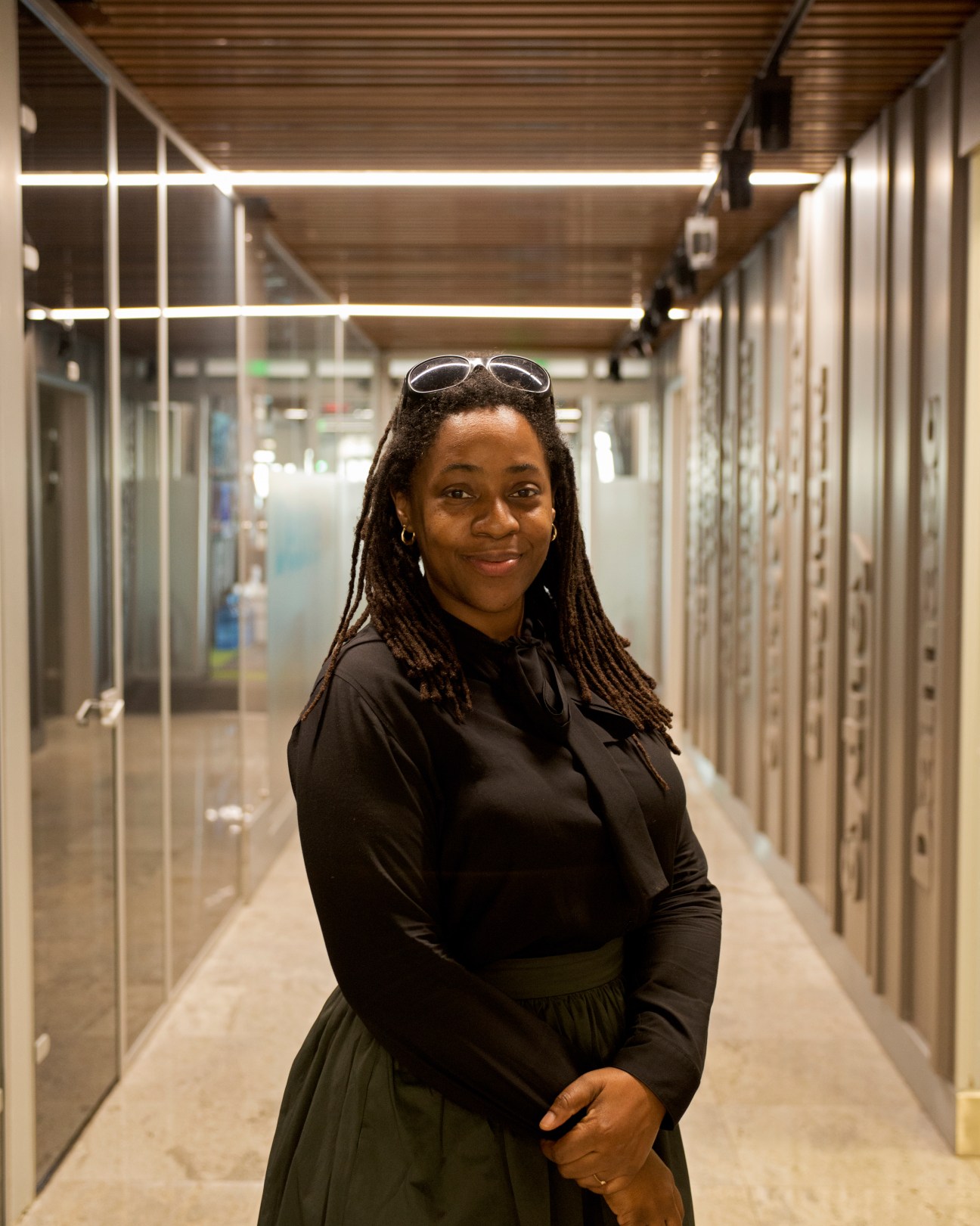 Cicily Scott stands in a modern hallway at TRT World offices, wearing a black top and olive skirt.