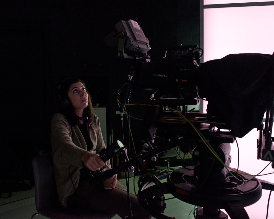 A Turkish camera operator operates professional broadcasting equipment in a dimly lit television studio.