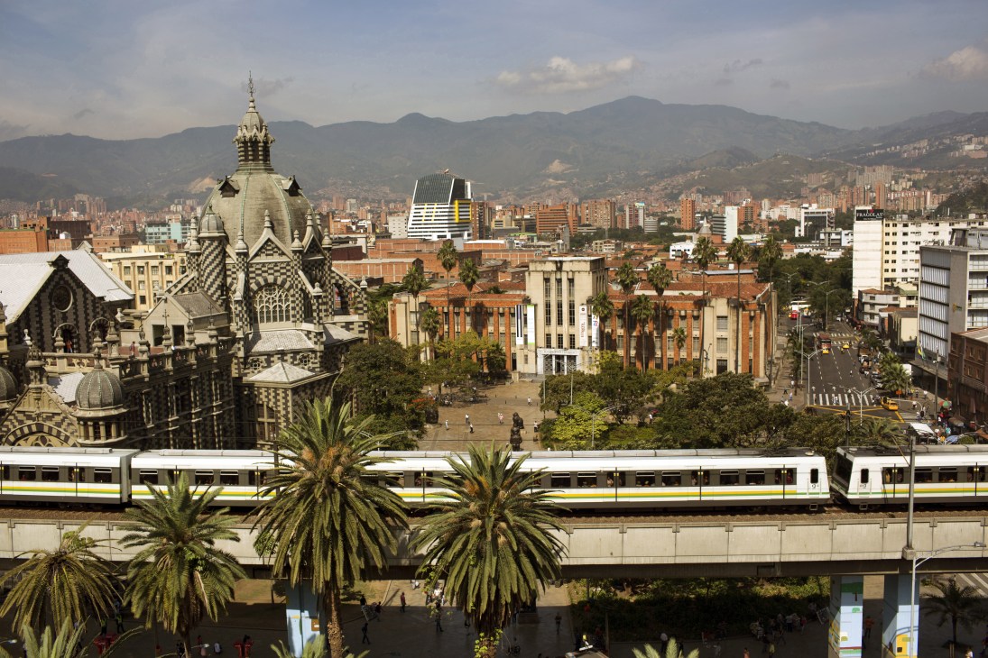 Medellín metro train passes ornate church with cityscape and mountains in background.