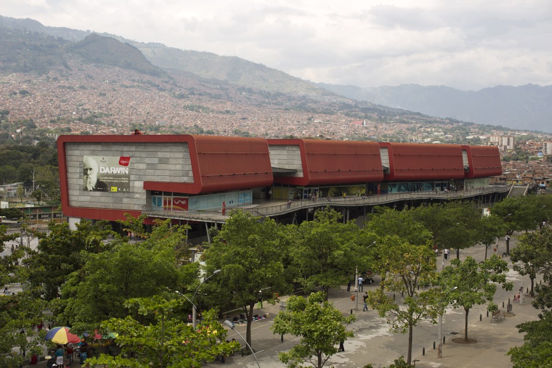 Parque Explora museum's distinctive red corrugated metal architecture overlooking Medellín's hillside slums and mountains.