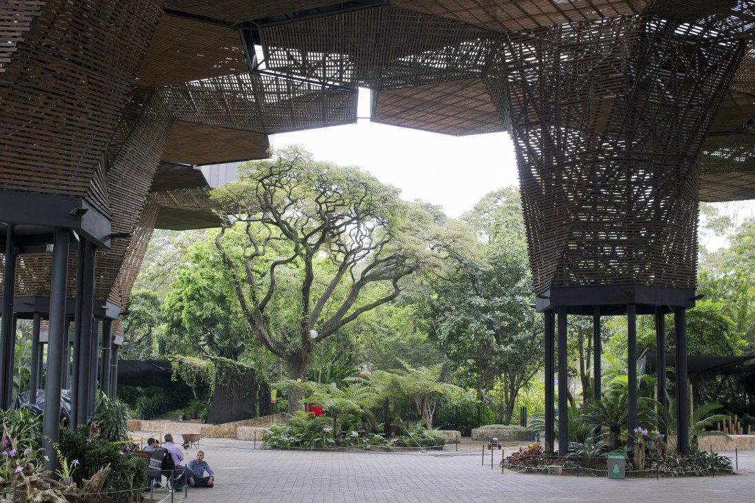 The Orquideorama botanical garden with distinctive black perforated metal canopy structures framing lush green trees and landscaped grounds.