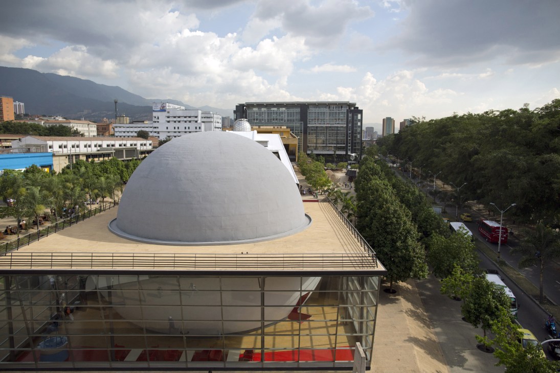A large white dome-shaped planetarium building with glass base structure sits on a plaza surrounded by modern city buildings and green vegetation.