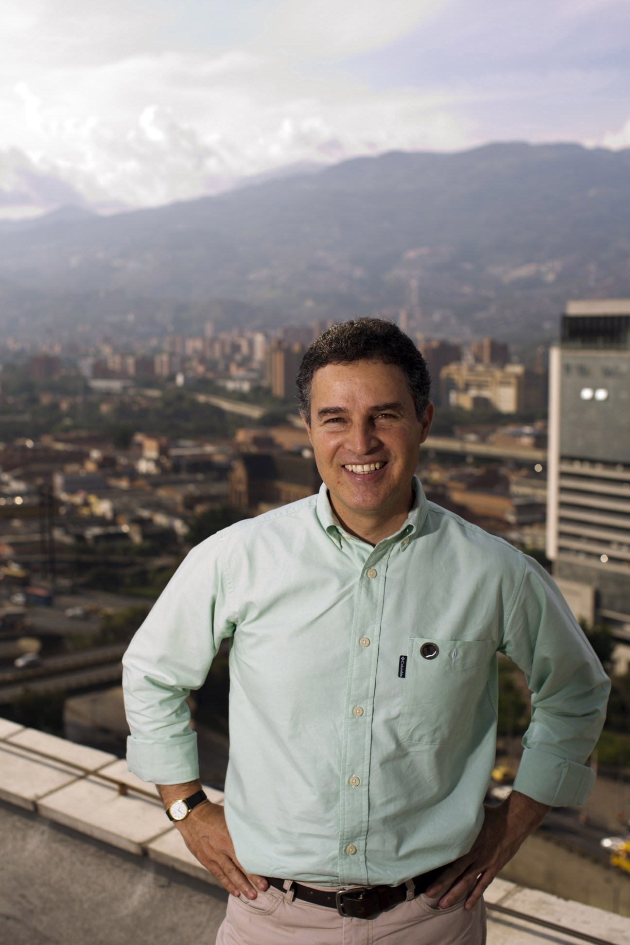 Mayor Aníbal Gaviria Correa smiling at a high-altitude vantage point overlooking Medellín's cityscape and surrounding mountains.
