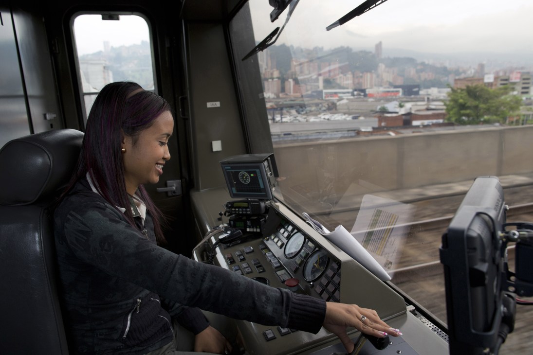 A female student operates the controls of a Medellín metro train from the cabin, with the city skyline visible through the window.
