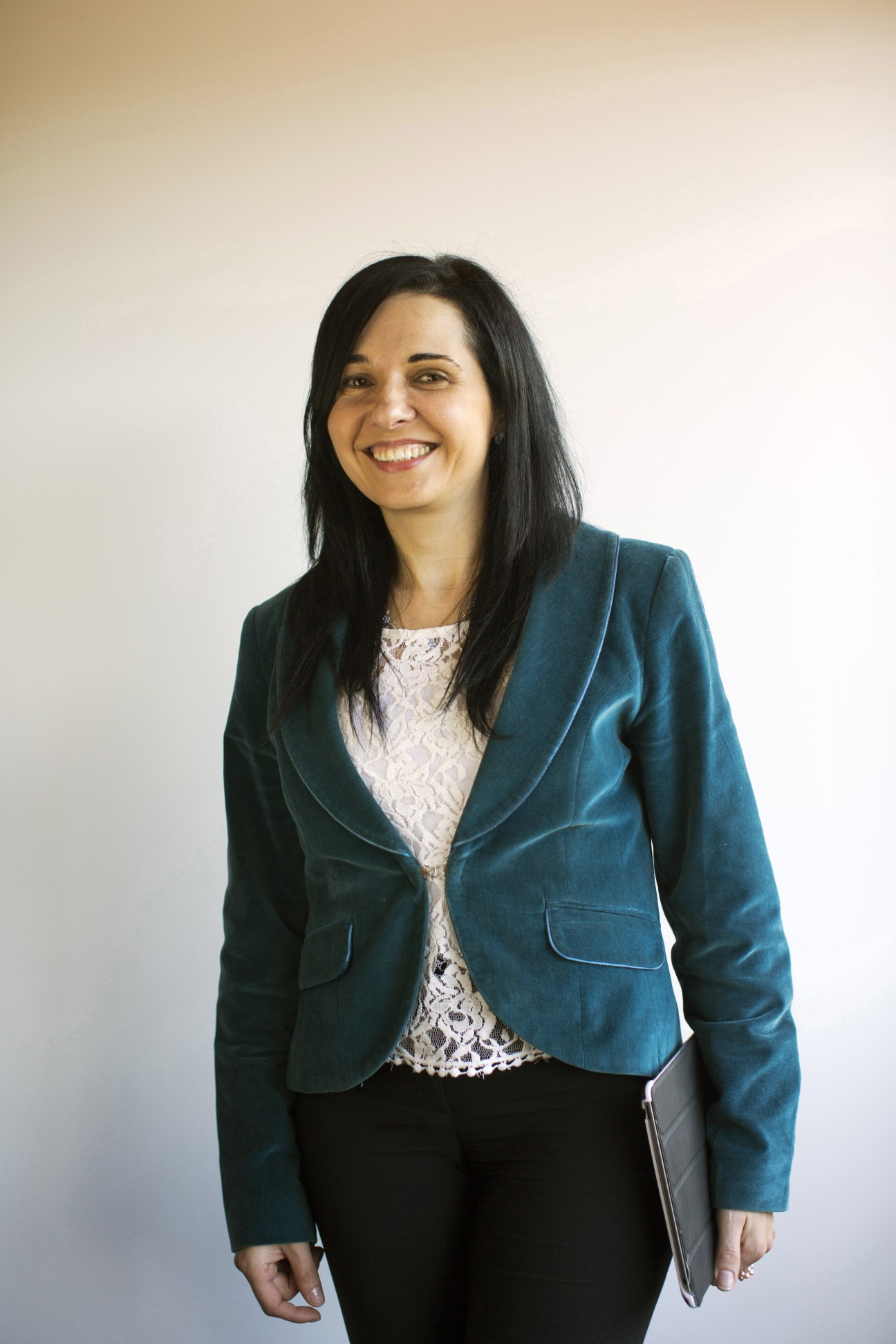 Woman in teal blazer and white lace top smiling at camera holding tablet.