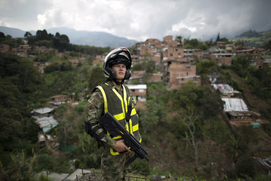Armed soldier in camouflage uniform and yellow safety vest standing on hillside overlooking Medellín slum settlement with brick buildings.