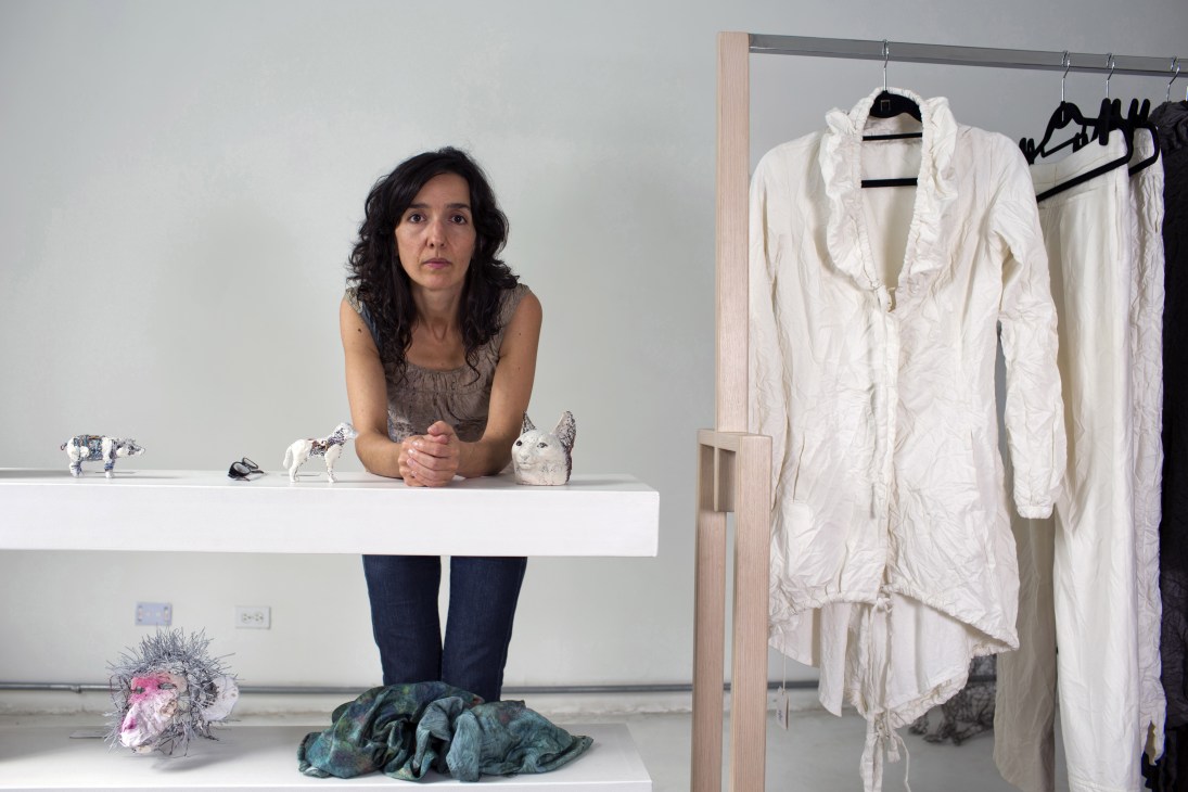 Fashion designer Juliana Correa sits at a white table with small sculptural pieces, wearing a grey sleeveless top and dark jeans, with white garments hanging on a rack behind her.