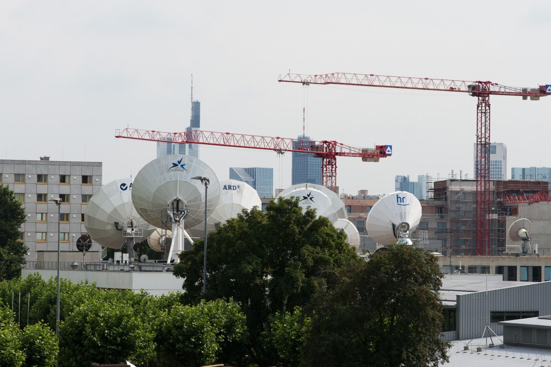 Large white satellite dishes on an HR broadcast facility rooftop with Frankfurt skyline and construction cranes in background.