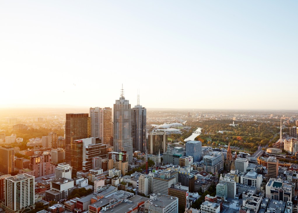 Aerial view of Melbourne's skyline with downtown skyscrapers, parks, and river visible at sunset.